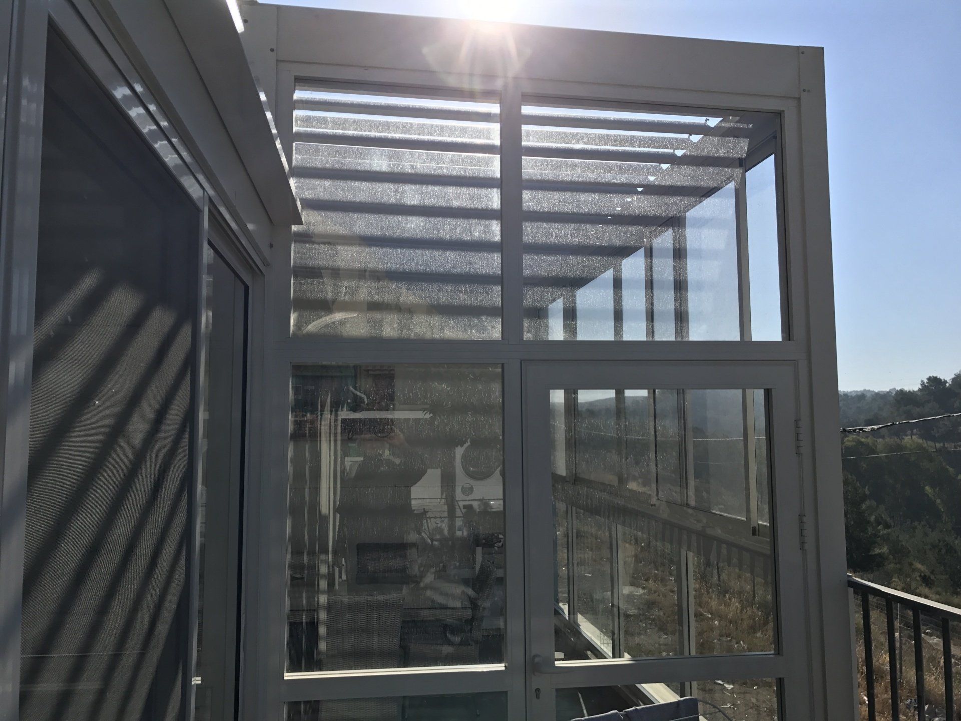 Sunlit glass-enclosed balcony with metal framing and a railing overlooking a distant landscape