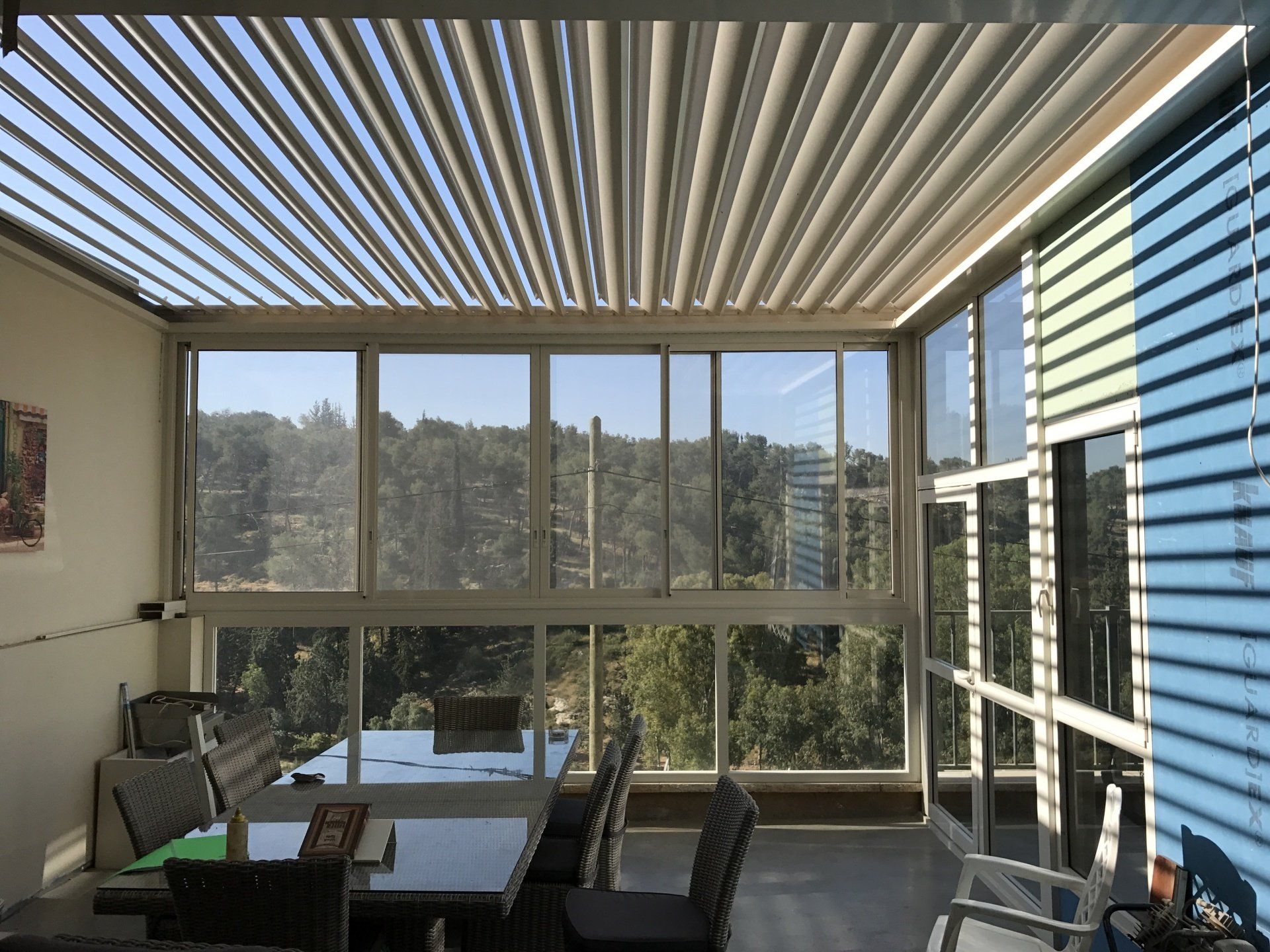 Sunlit modern patio with striped pergola, glass walls, chairs, and mountain view