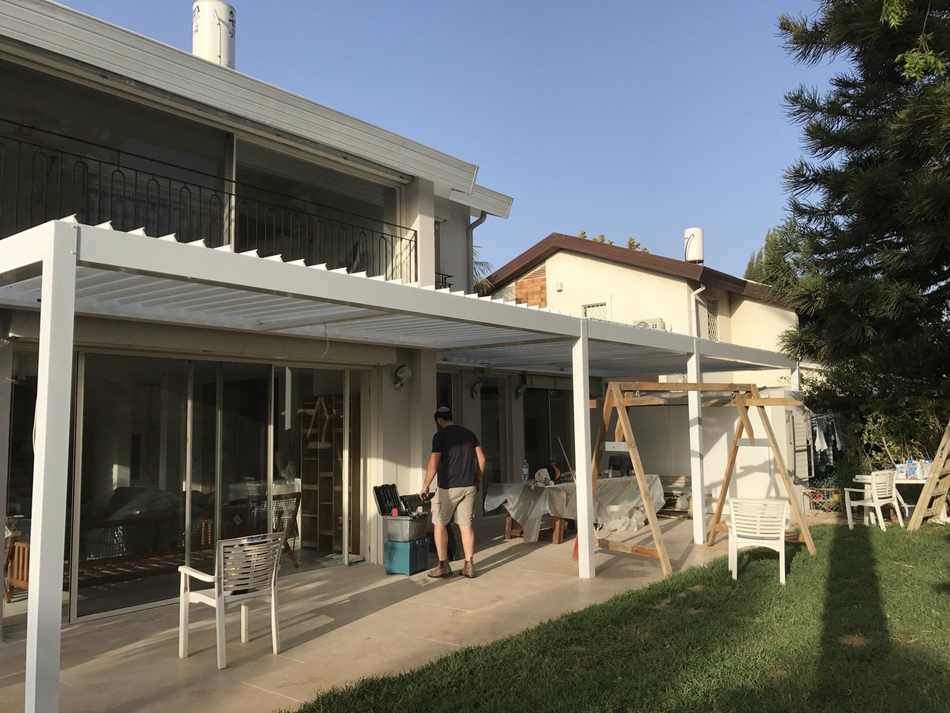 Two people working on a patio beside a modern house, with a white pergola and lawn in sunlight.