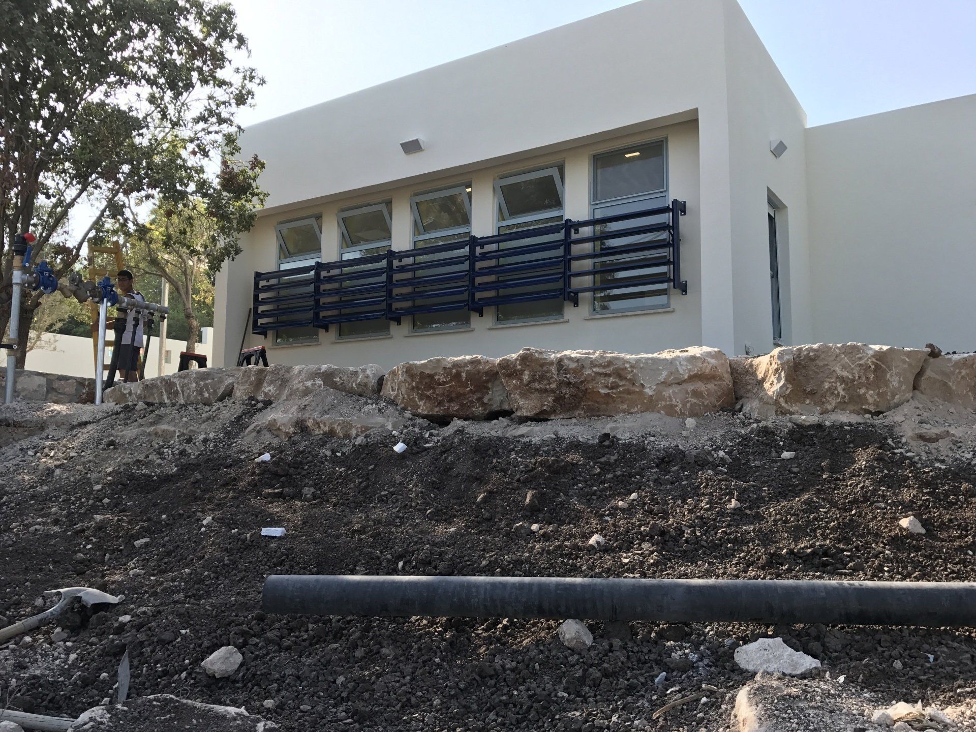 Modern white house with dark balcony railings behind a rocky dirt slope and trees