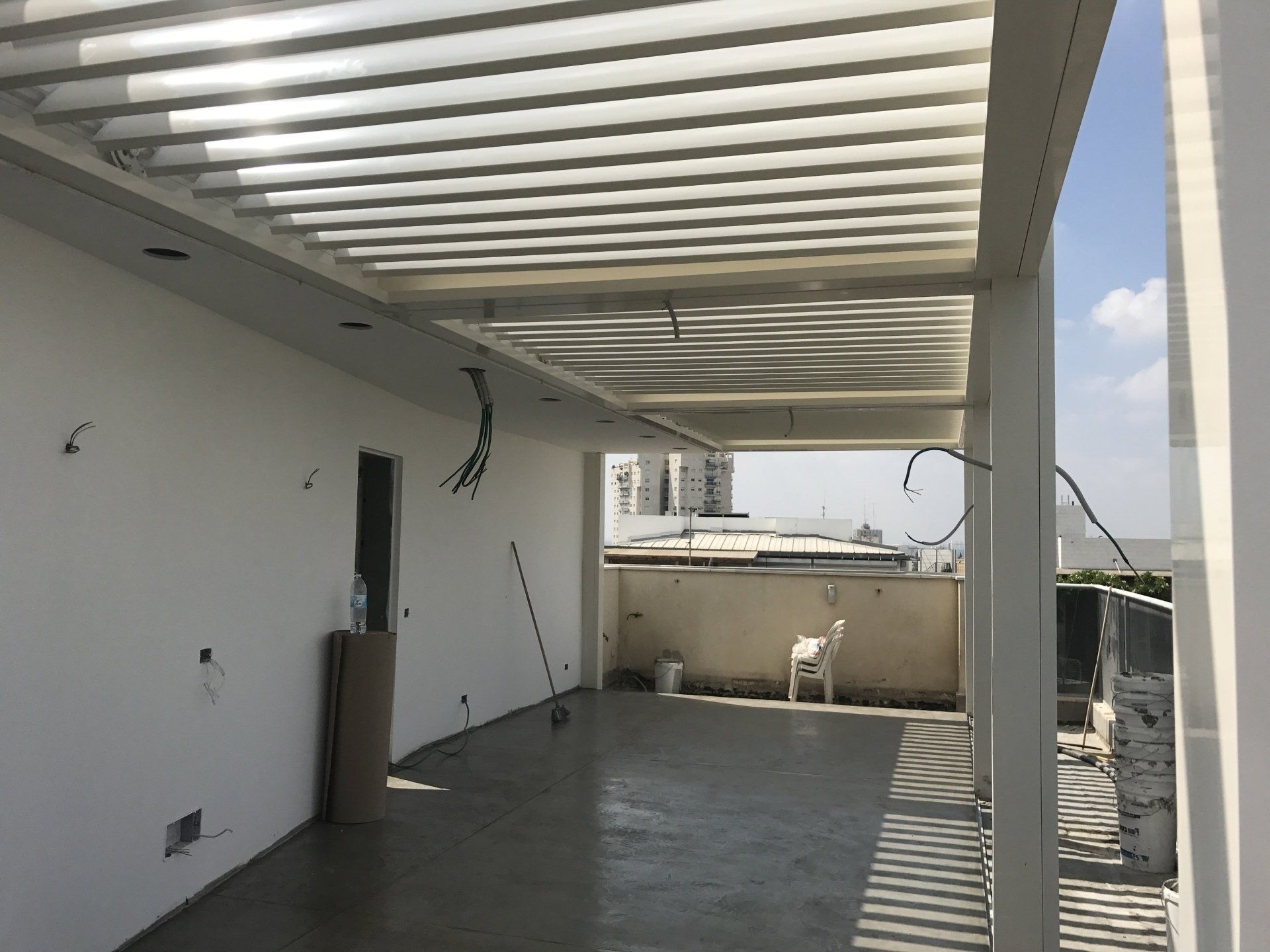 Empty covered rooftop terrace with white pergola slats, concrete floor, and city view beyond the wall