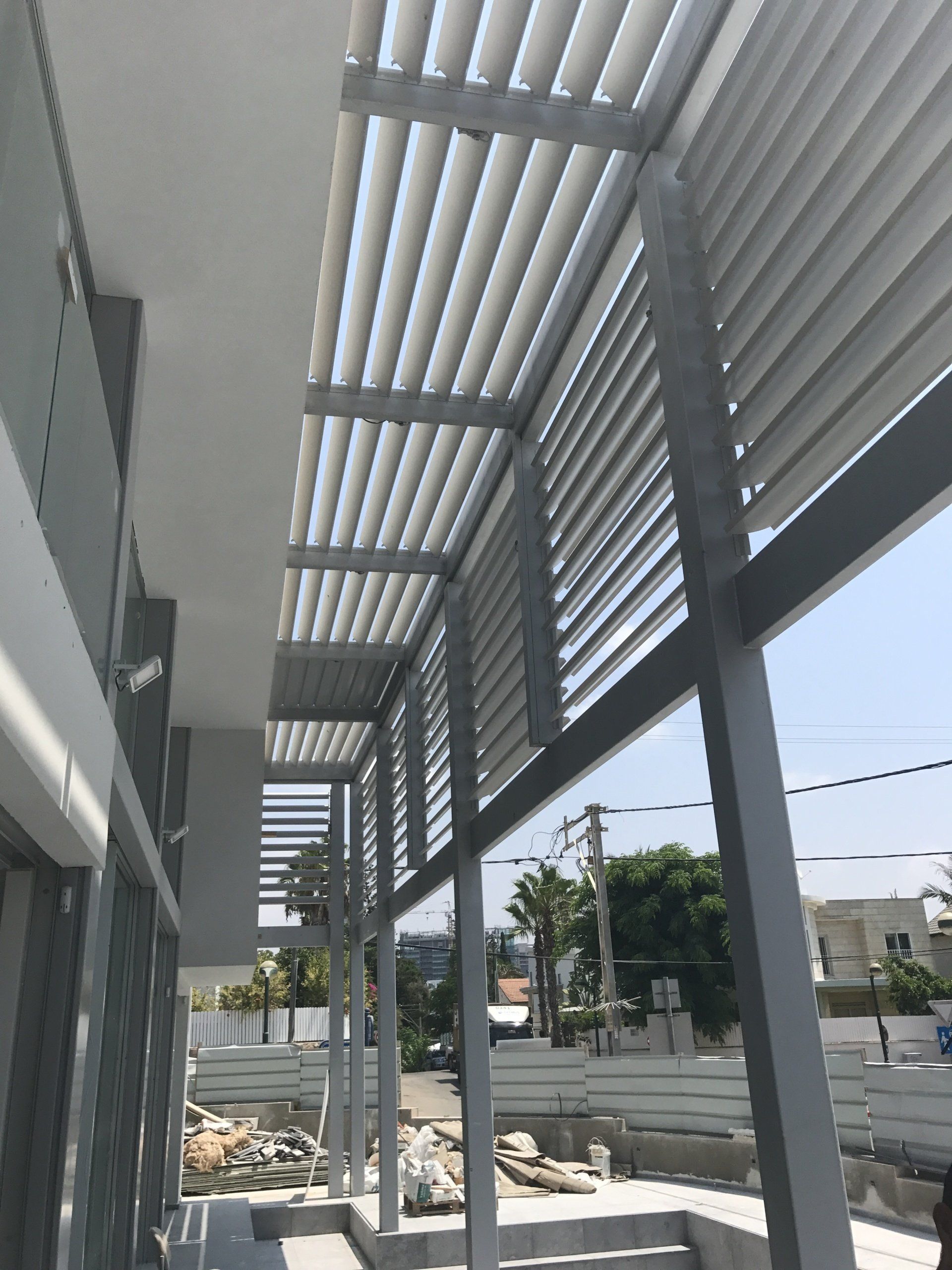 Modern building walkway with gray metal columns and overhead slatted shade casting striped shadows.