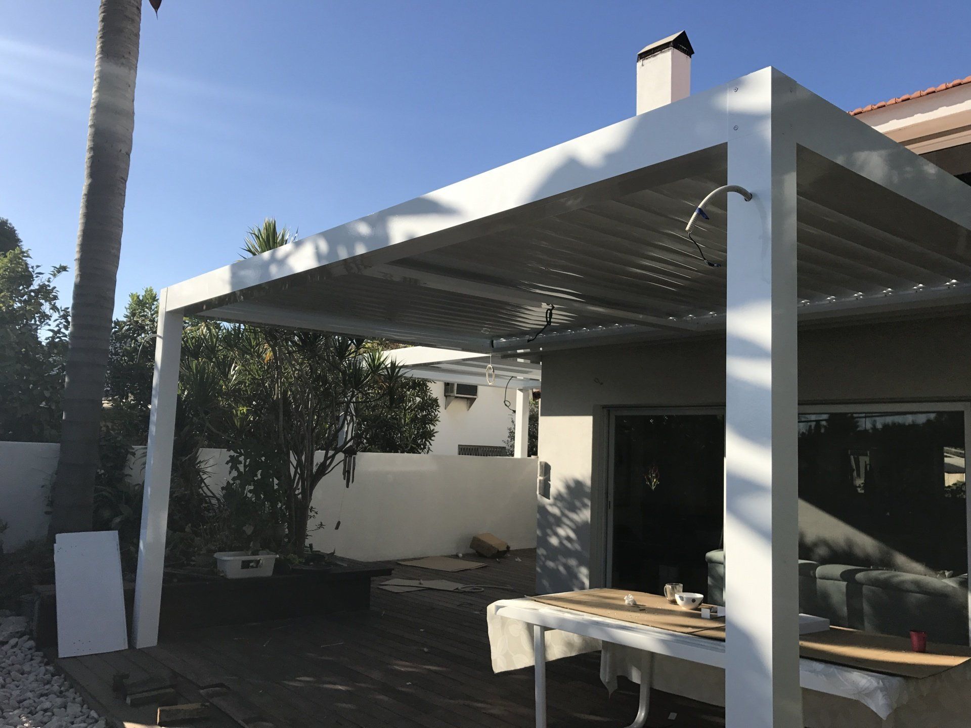 Patio with a white pergola roof, outdoor table, and potted plants beside a house in sunlight