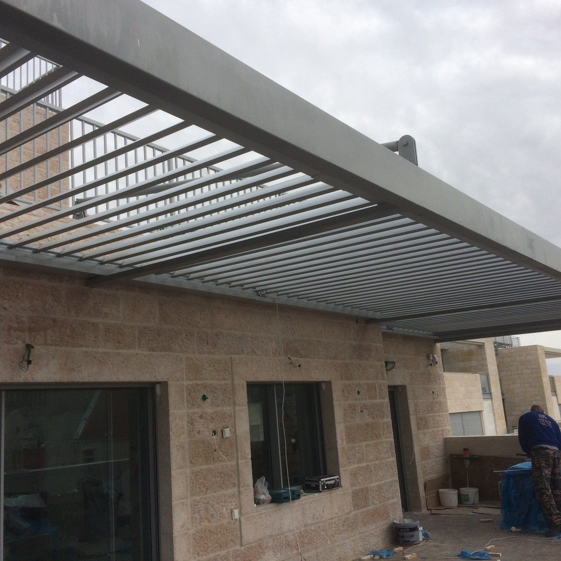 Stone house under construction with a metal pergola roof and a worker at the right side