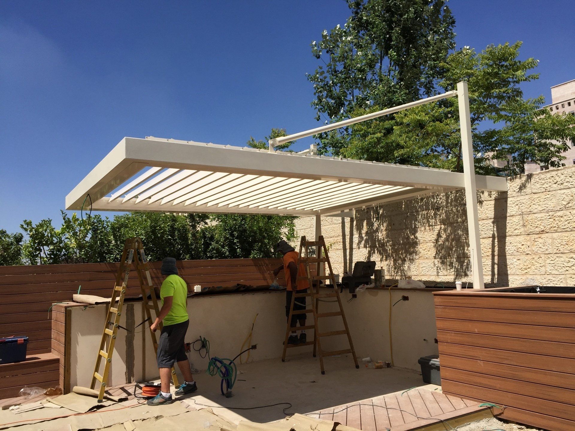 Two workers building a white patio pergola beside a beige wall under a blue sky