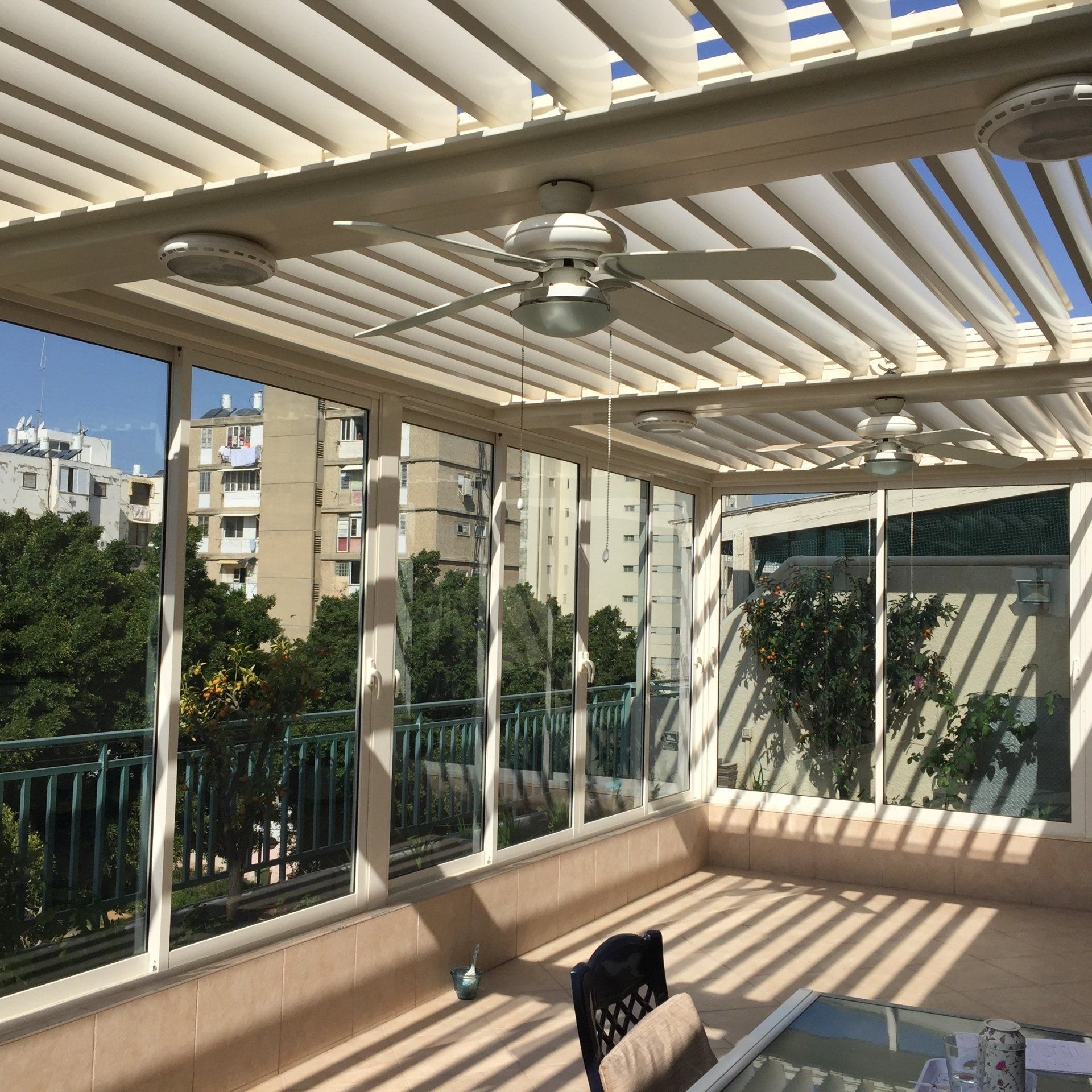 Sunny balcony with white pergola, ceiling fan, city buildings, and railing shadows