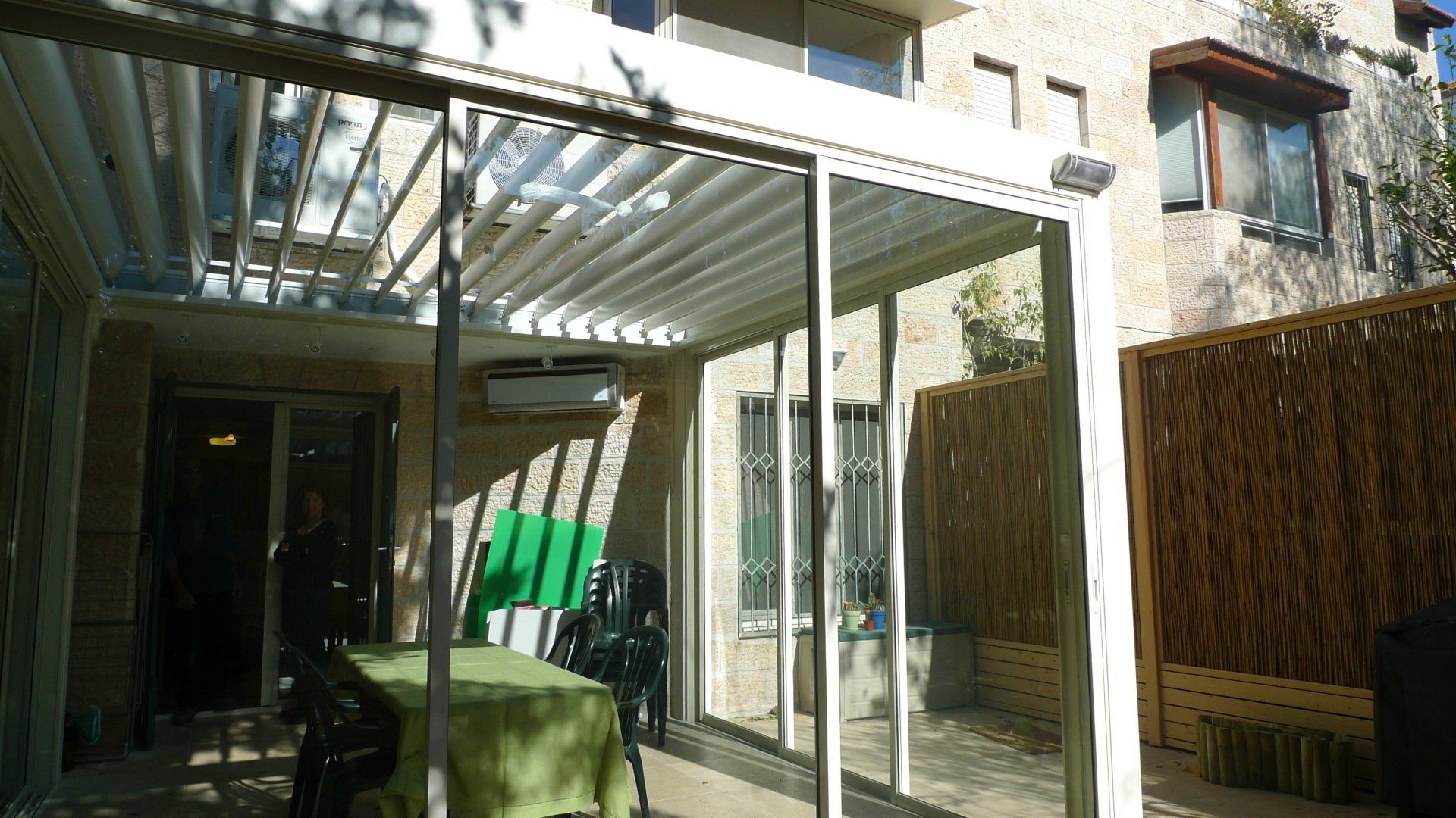 Backyard patio with a white pergola, glass roof, green chairs, and a wooden fence beside a house.