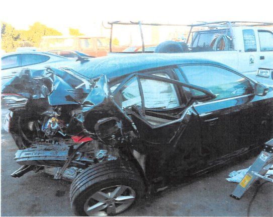 Heavily damaged black sedan with extensive front-end collision damage and deployed airbags in an outdoor setting.