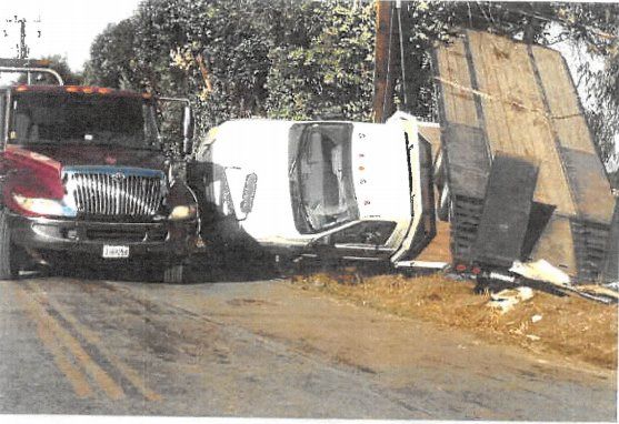 A car is overturned and crashed beside a flatbed trailer and a large truck. The vehicles are on the side of a road.