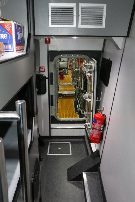Narrow industrial stairwell with metal steps, vents, and a red fire extinguisher beside a glass door.