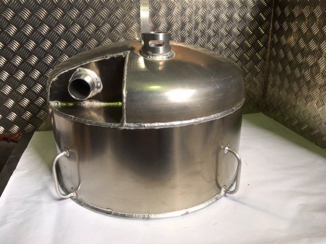 Stainless steel tank with side spout and handles on a white surface against a patterned metal wall.