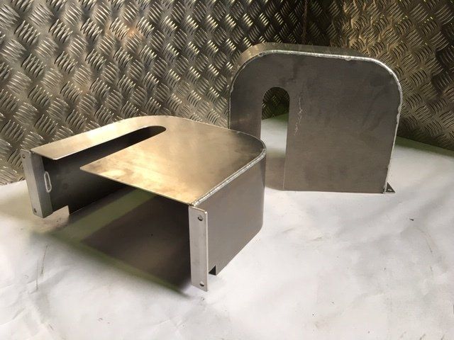 Two stainless steel metal parts on a workbench, one bent into a U-shape with a cutout.