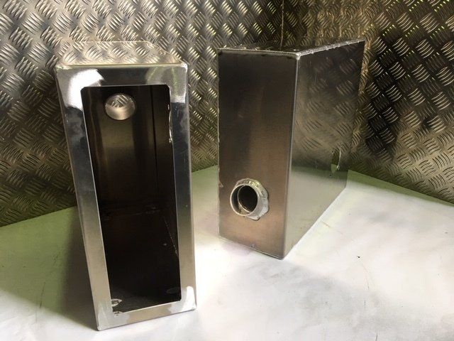 Two stainless steel rectangular enclosures, one open with a bolt inside, on a textured metal surface.