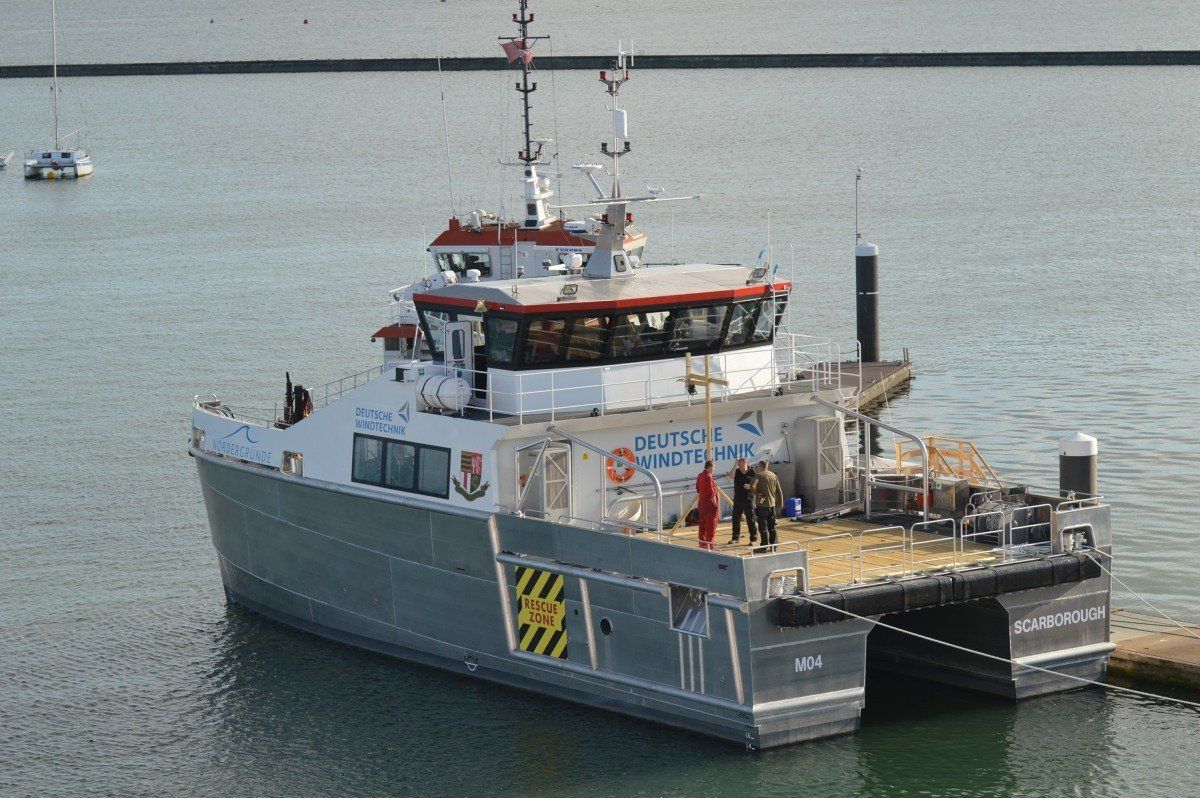 Large gray catamaran ferry on calm water with people standing on its deck