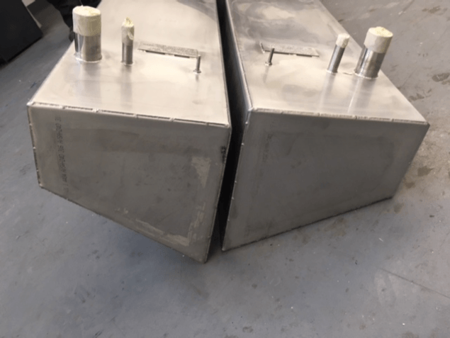 Two metal wedge-shaped containers with bolts and white caps on a gray floor