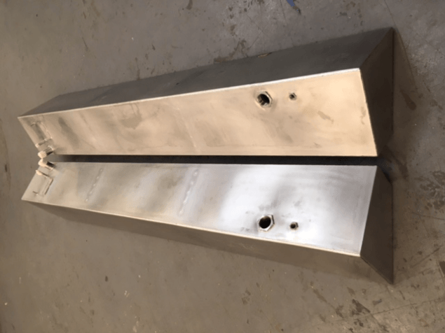 Two brushed metal wedge-shaped panels with holes, lying on a concrete floor.