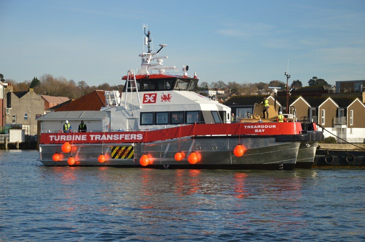 Red and white ferry “Turbine Transfer” on calm water near houses and a dock with orange buoys.