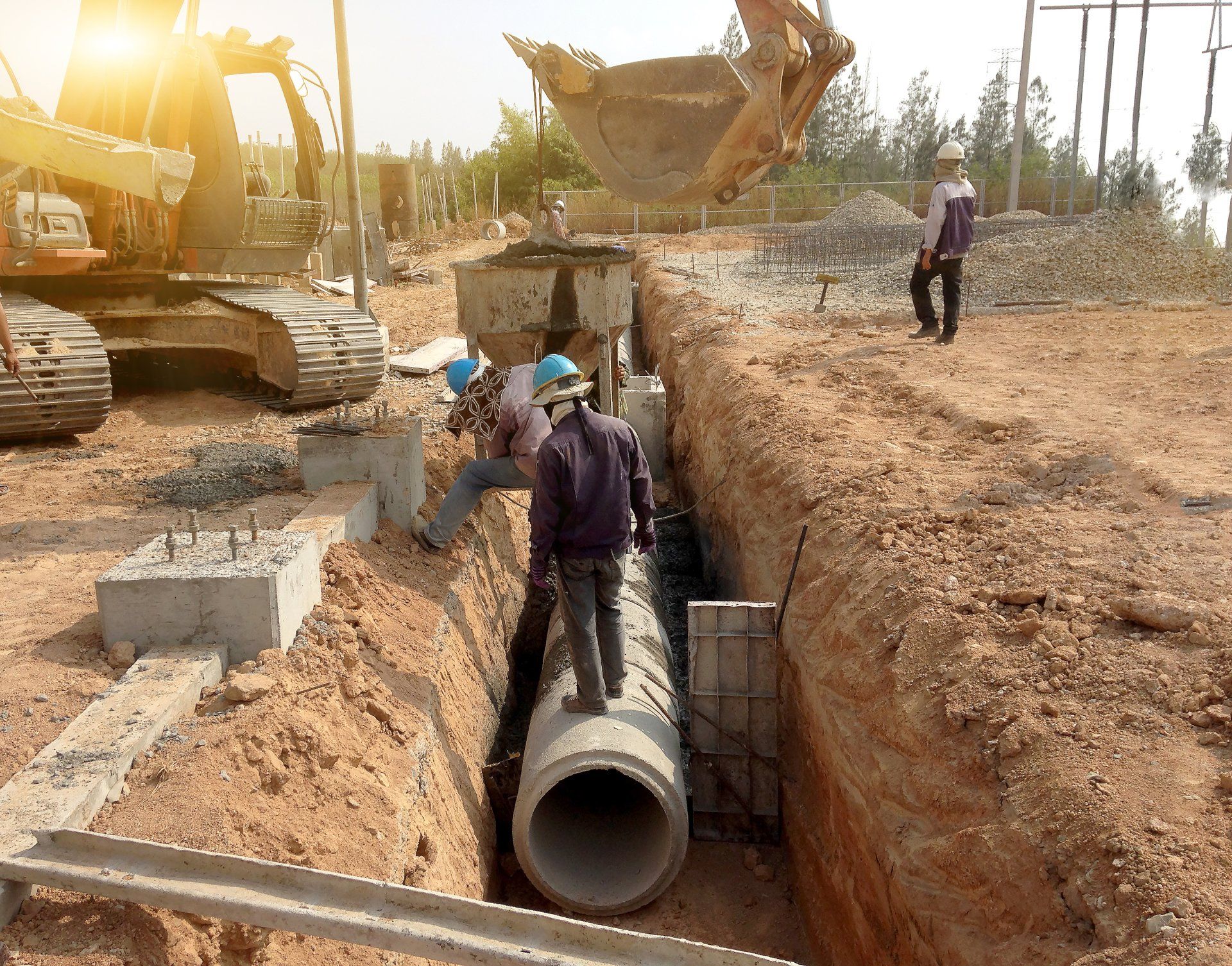 Construction workers laying a large drainage pipe in a trench beside an excavator on a dirt site.