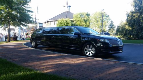Black limousine parked on a brick sidewalk near a green lawn and a building with a steeple.