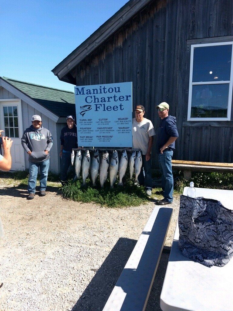 People posing beside sign for Manitou Charter Fishing at a dark blue building with fish displayed underneath.