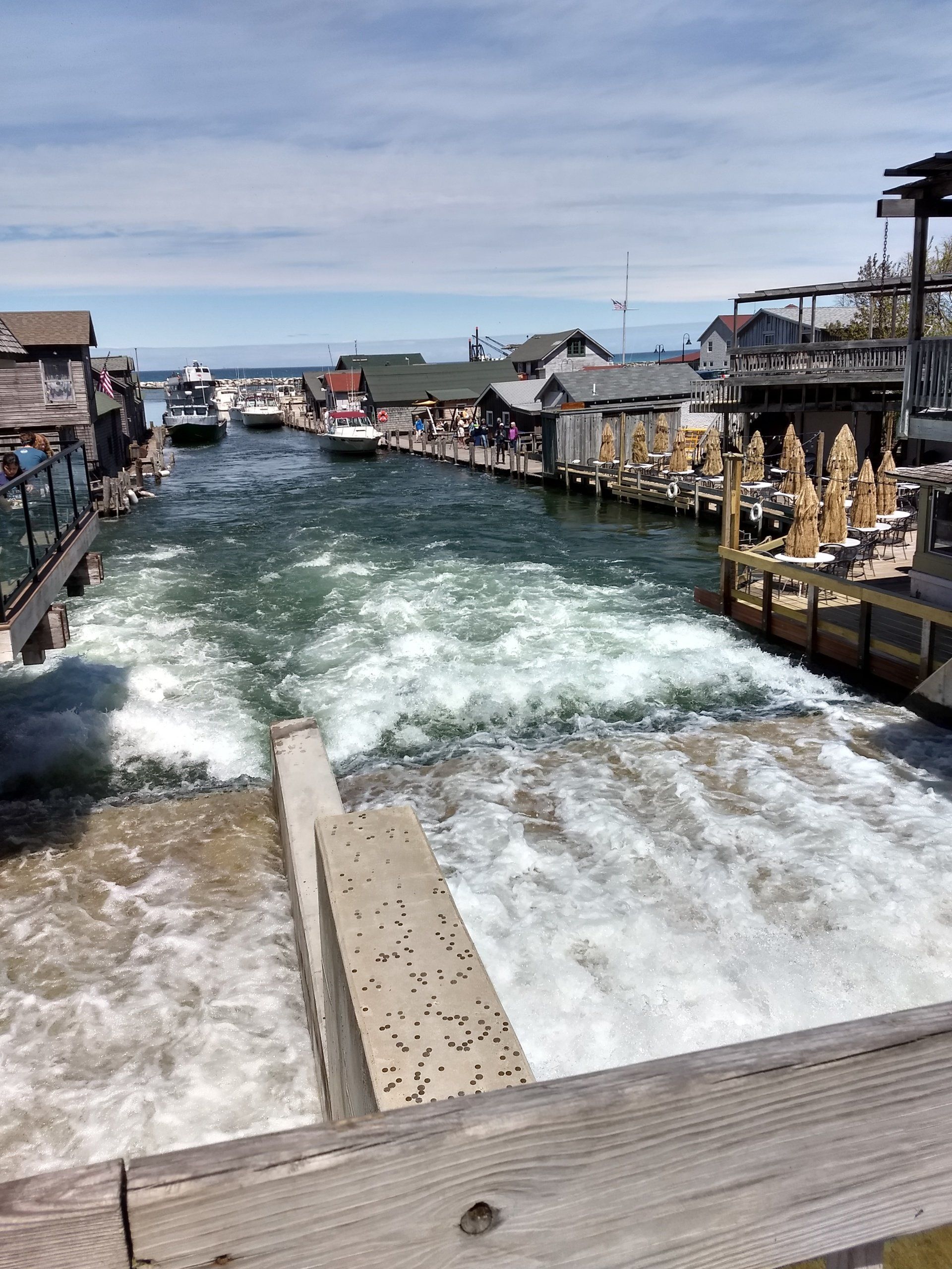 Canal lock with churning water, lined by boats and wooden docks under a bright sky