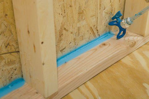 Applying blue spray foam insulation to a wooden wall frame.