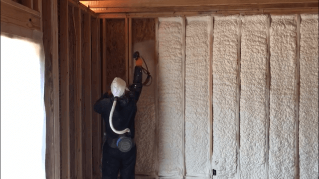 Person spraying insulation foam onto a wooden wall, wearing protective gear.