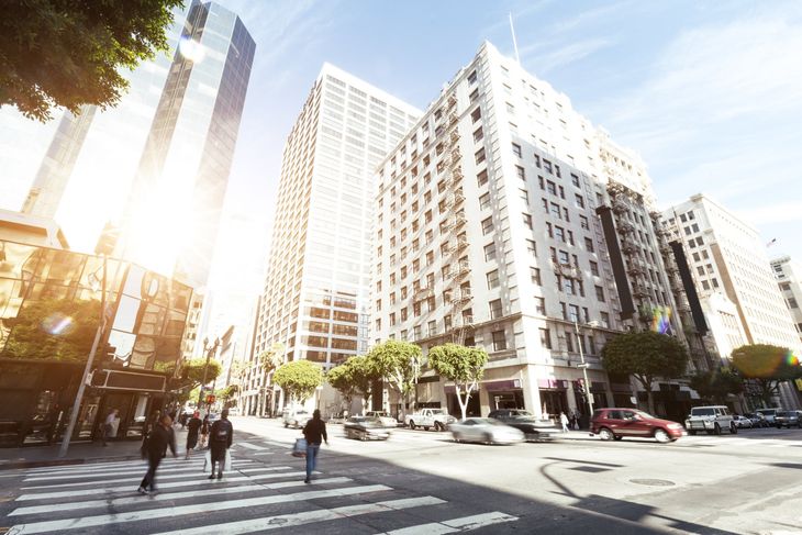 Sunlit city street with pedestrians, tall buildings, and a crosswalk at an urban intersection