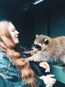 Woman smiling, playfully interacting with a raccoon inside a dark enclosure.