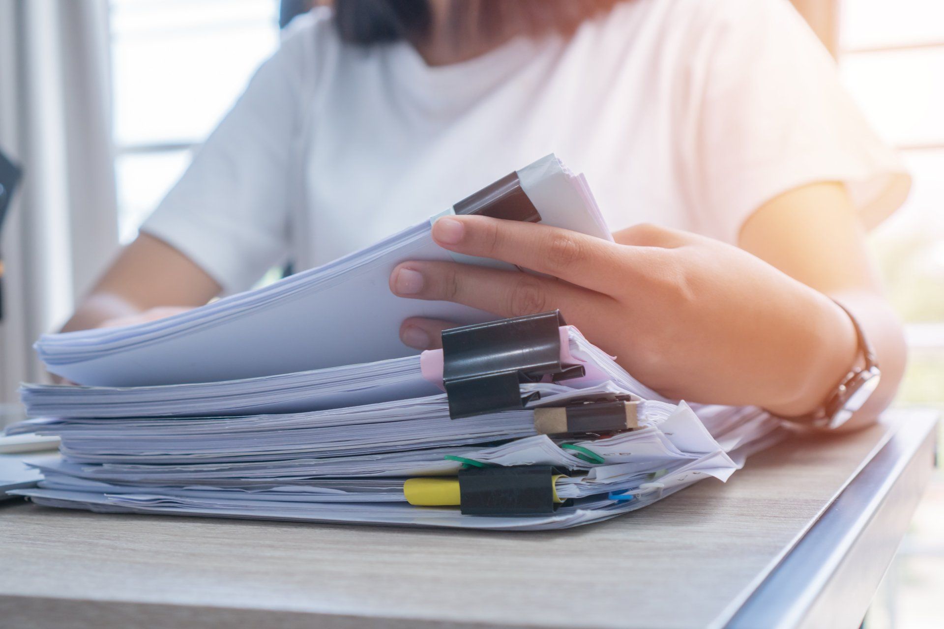 Person holding a stack of papers and notebooks at a desk, with clips and a pen visible