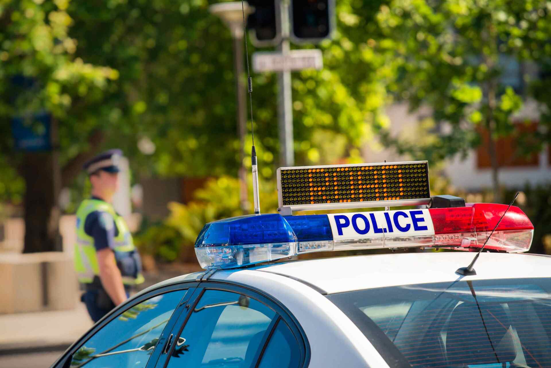 Police car with flashing light bar parked on a sunlit street, officer blurred in the background