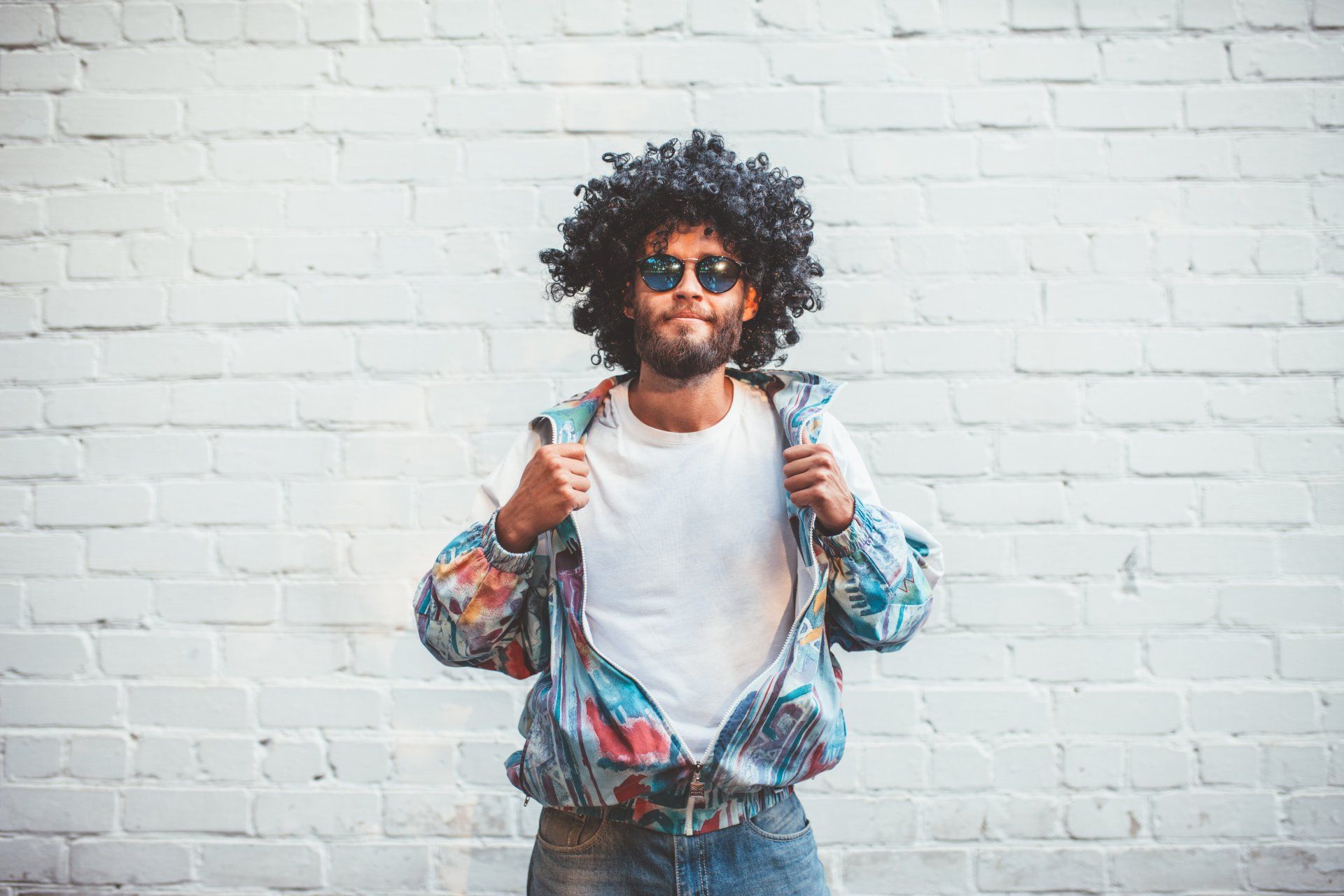 A man with an afro and sunglasses is standing in front of a white brick wall.