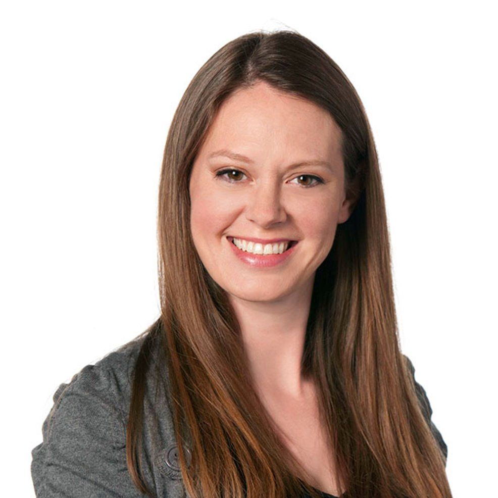 Woman with long brown hair smiles, wearing a gray jacket, against a white background.