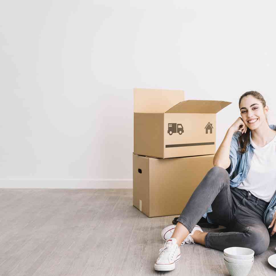 Woman smiling, sitting on floor next to moving boxes, leaning on hand.