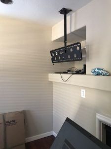TV mounted on a ceiling arm, above a mantel. Wall is light tan. Brown floor.