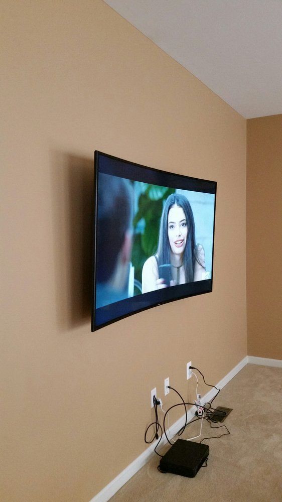 A curved TV mounted on a tan wall, showing a woman on screen. Electrical cords and a black box sit below.