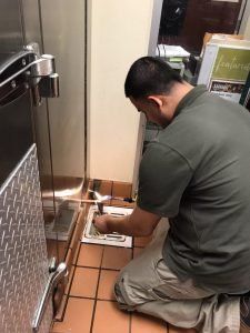 A person kneels on a tile floor, working on a door frame in a commercial kitchen.