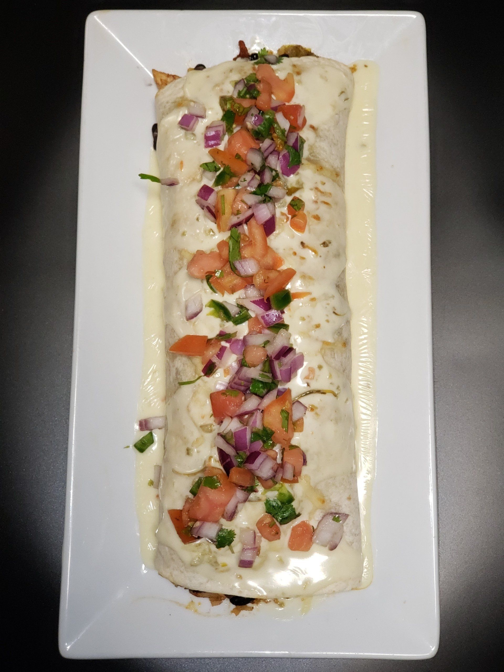 Burrito covered in creamy sauce, topped with chopped tomatoes, red onion, and cilantro on a white rectangular plate.
