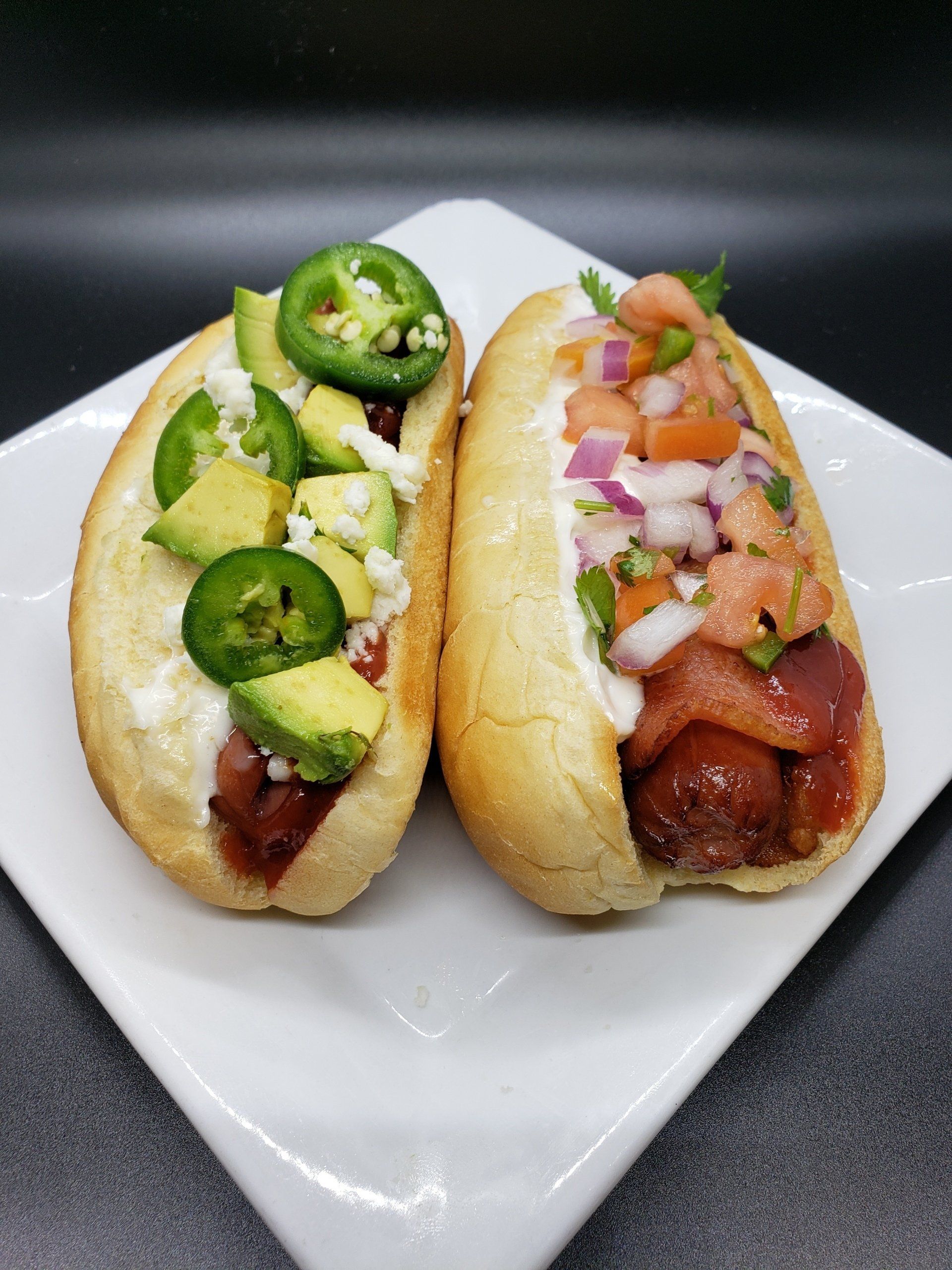 Two gourmet hot dogs on a white plate; one with avocado, jalapeños, and cheese; one with pico de gallo.