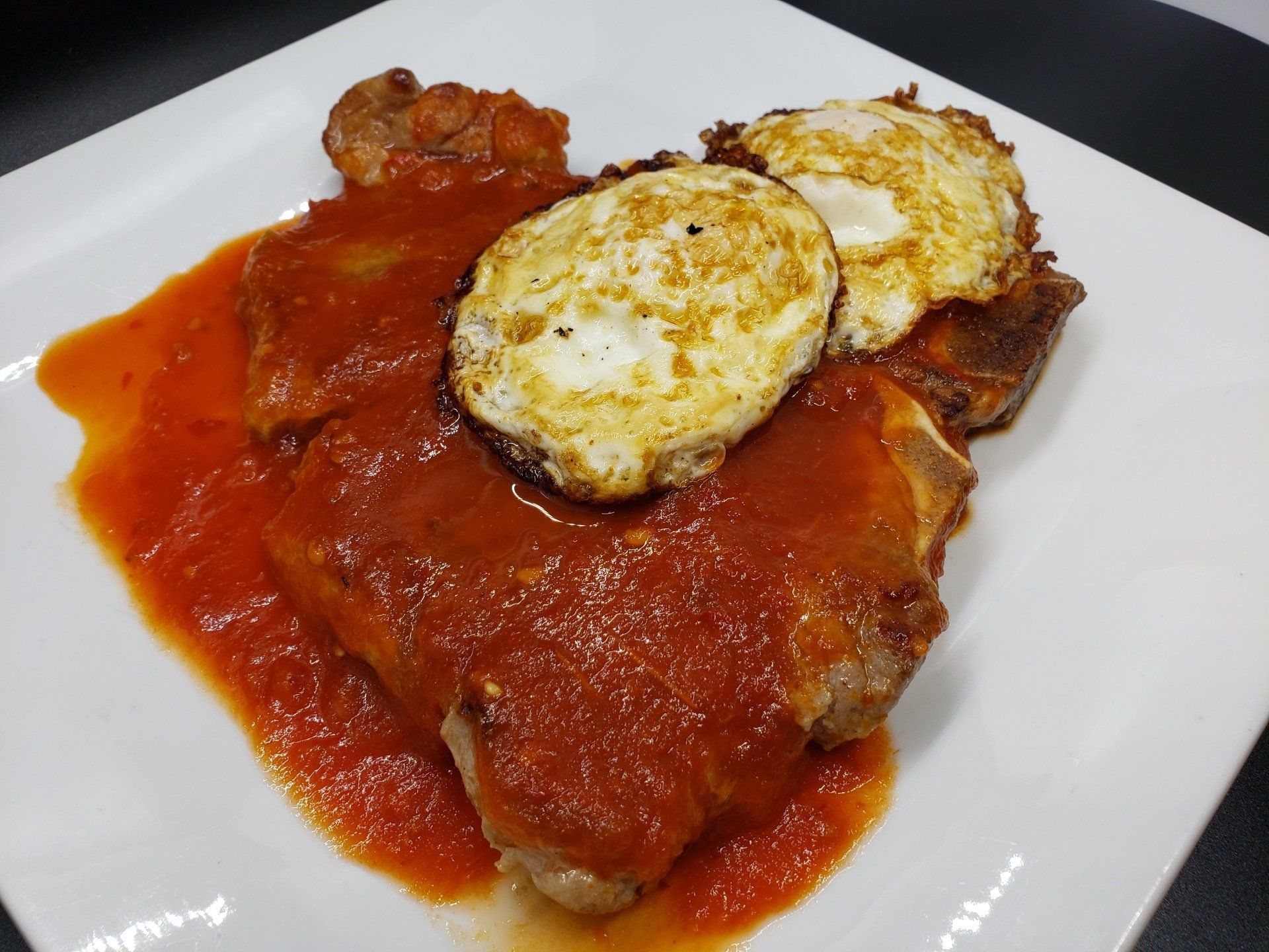 Pork chops in tomato sauce topped with two fried eggs on a white plate.