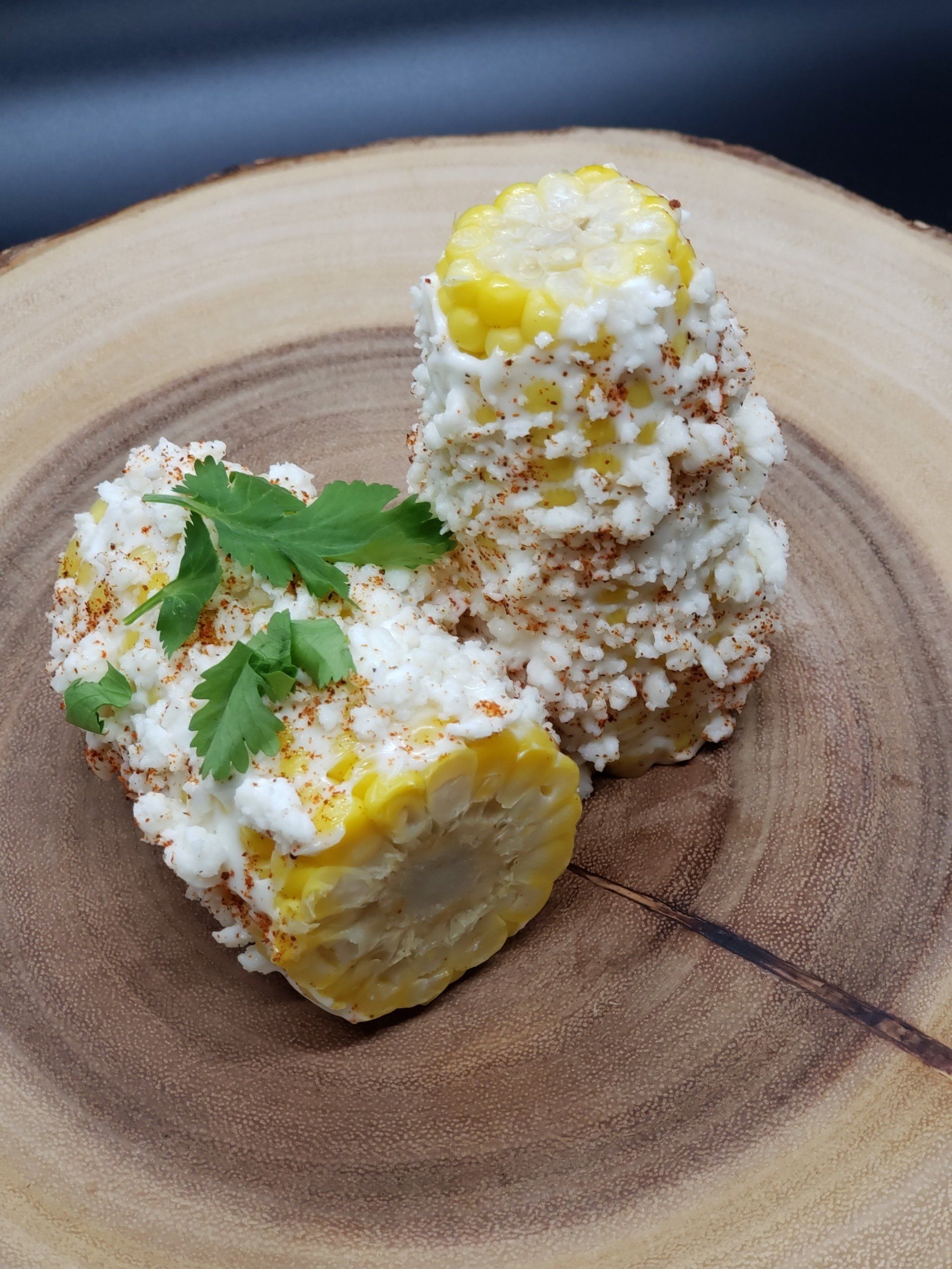 Grilled corn on the cob, stacked, covered in white cheese, paprika, and cilantro on a wooden platter.