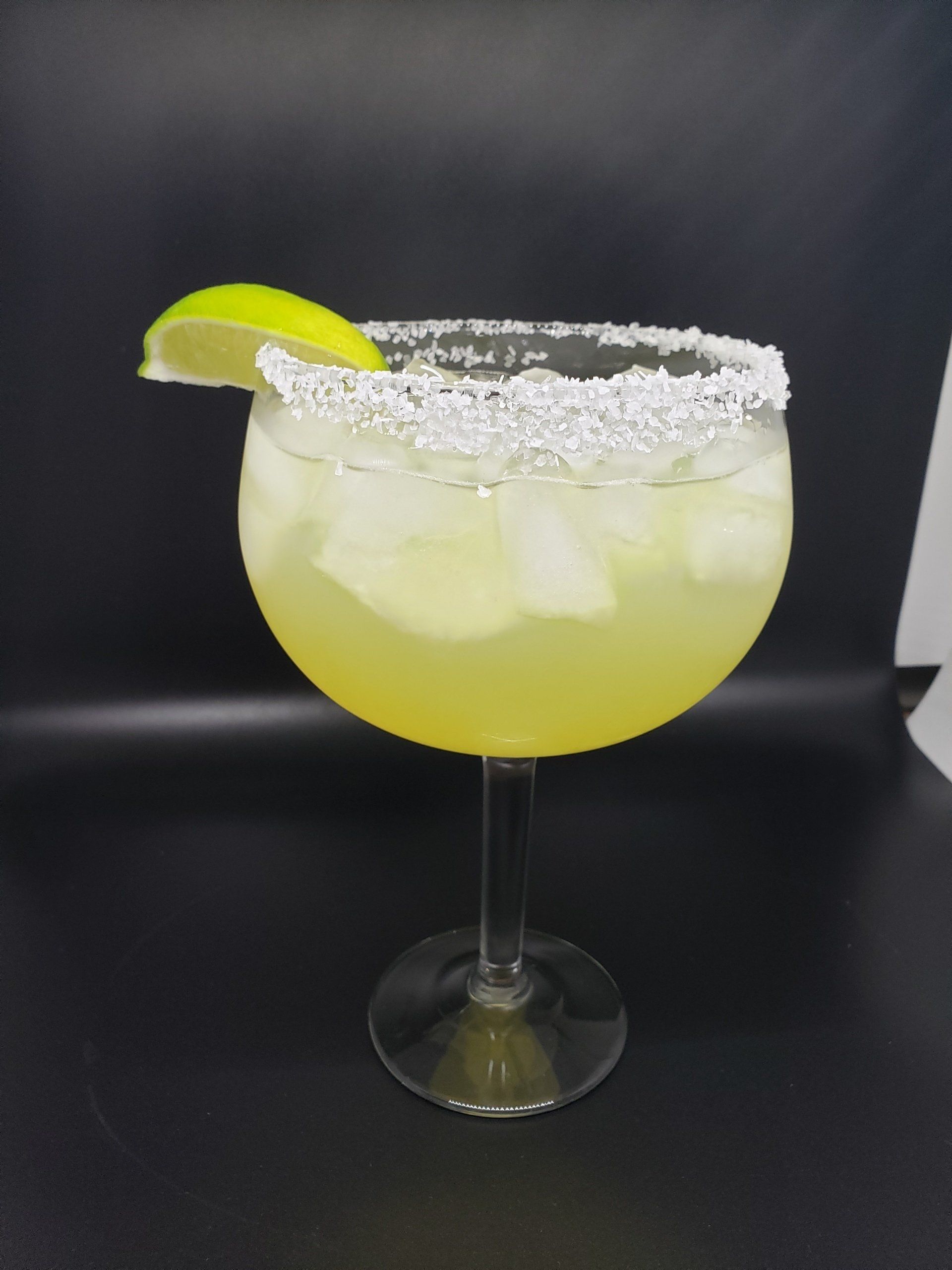 Margarita cocktail in a balloon glass, salt rim, lime wedge garnish, ice, against a black background.