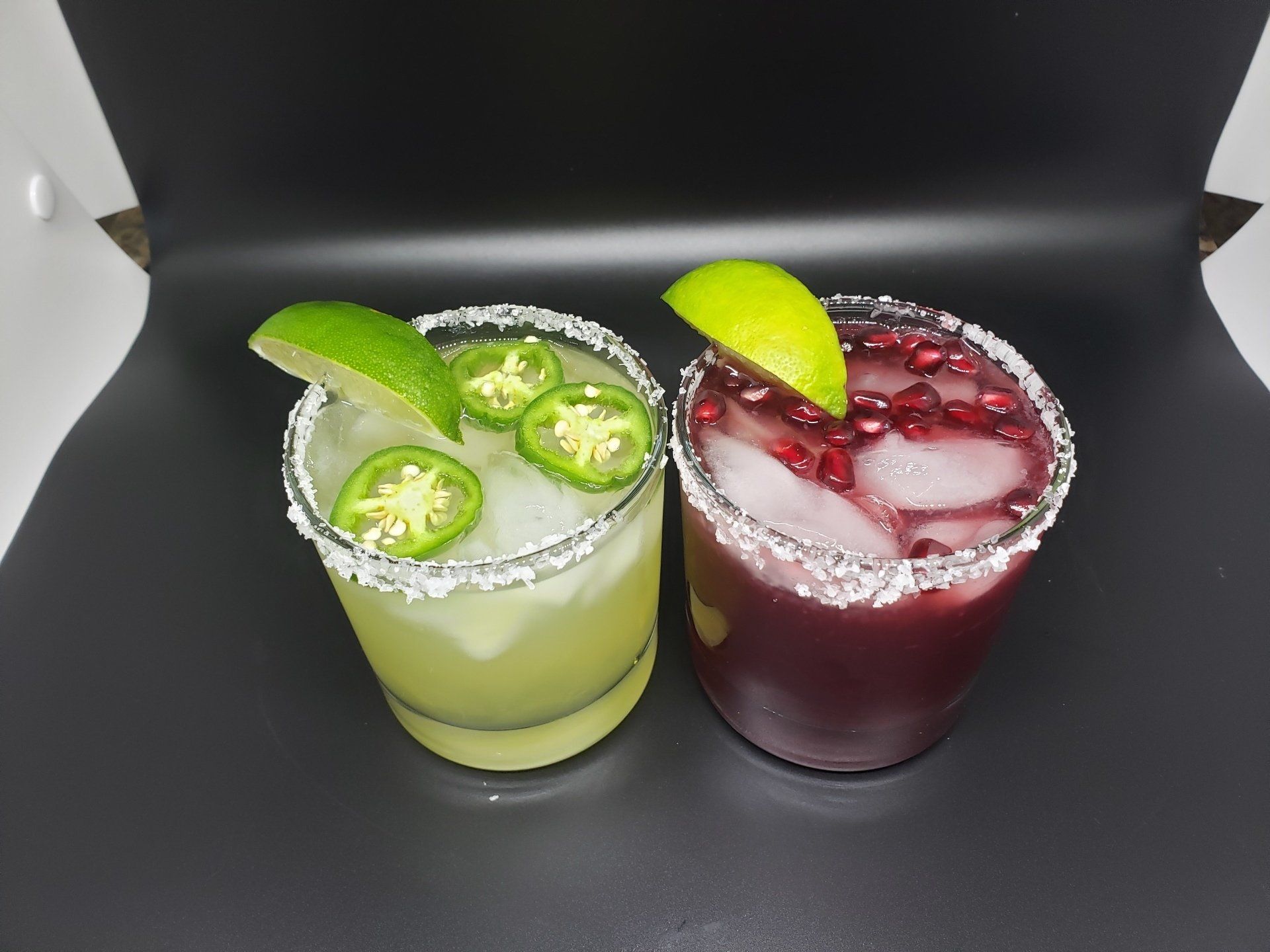Two cocktails in glasses, one green, one red, with lime and jalapeno garnishes on a dark surface.