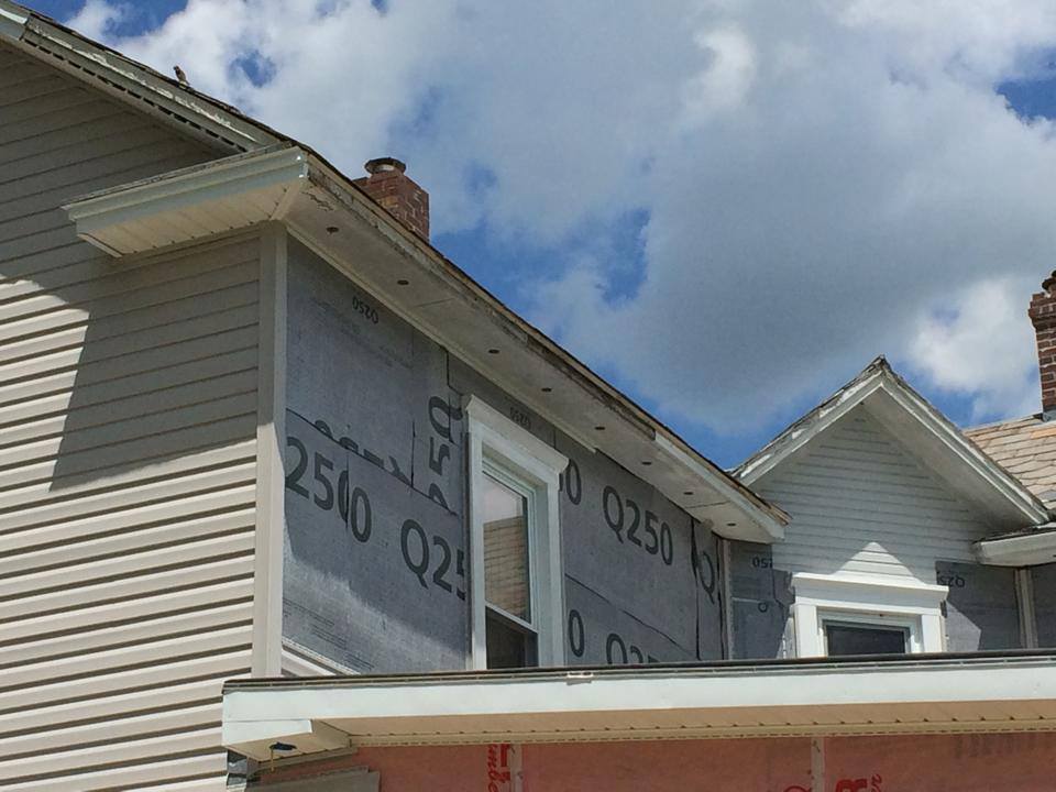House exterior under repair with white siding, housewrap, and a partly visible chimney against a cloudy sky