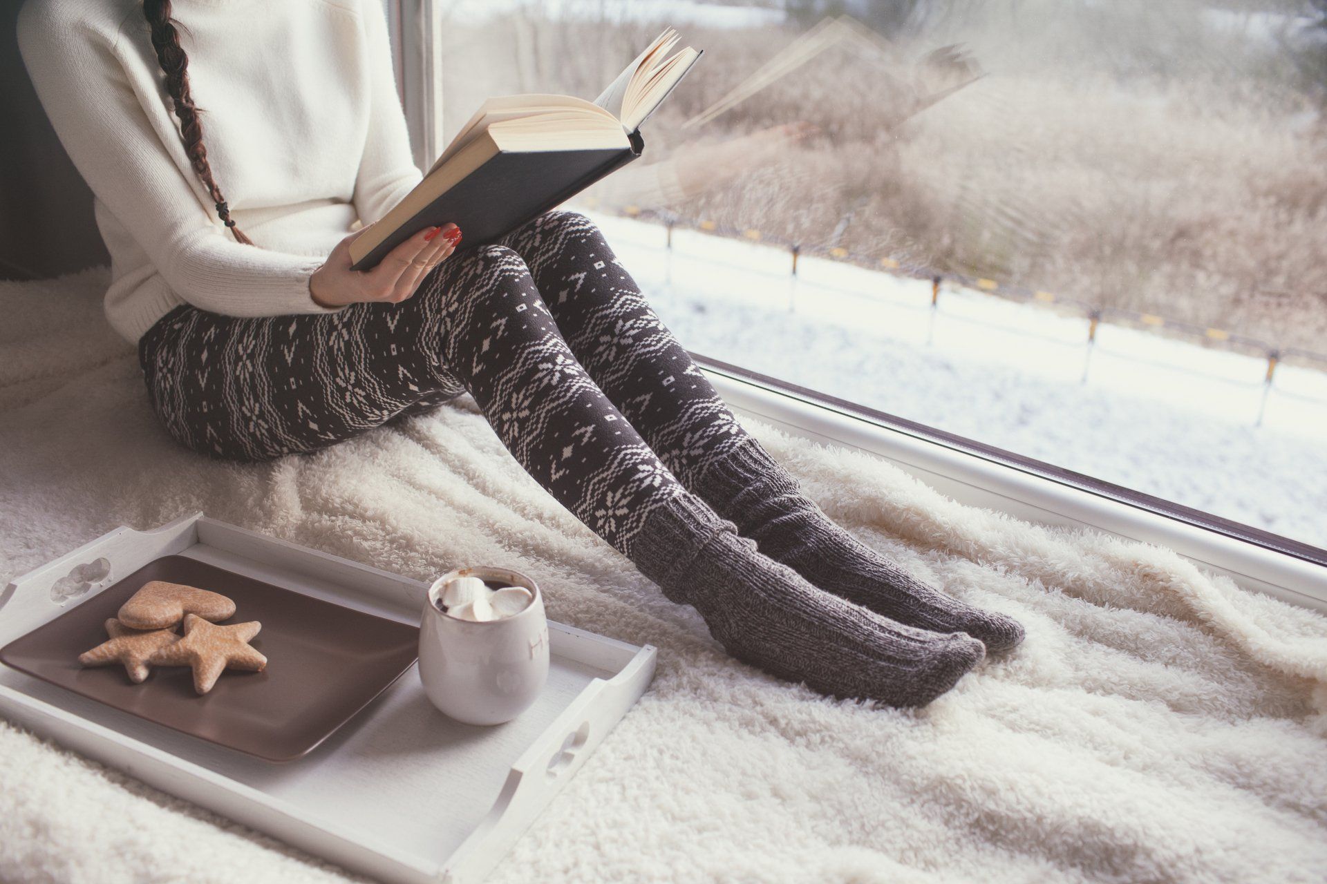 Image of a woman reading a book in front of a large window wearing winter clothes
