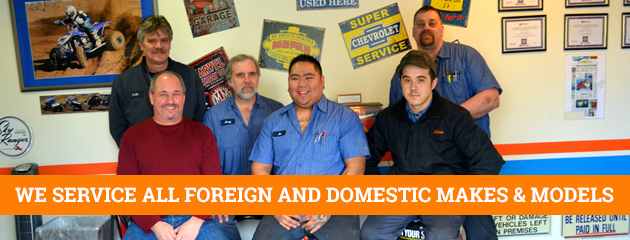 Five smiling auto shop staff posing in a garage with “We service all foreign and domestic makes & models” banner