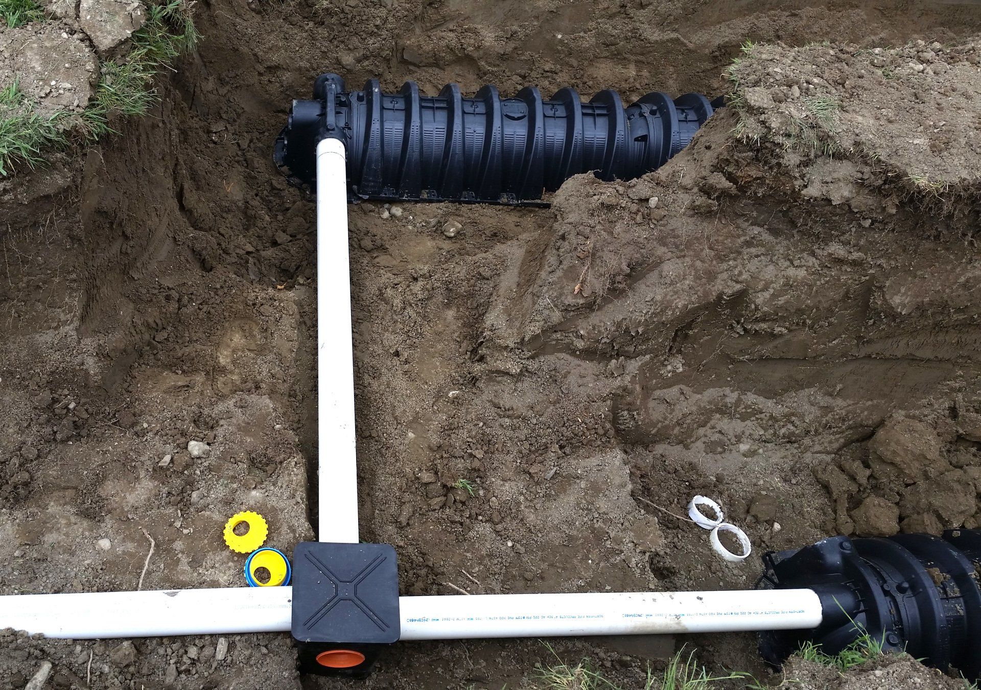 Black plastic septic drainage chambers connected by white PVC pipes in an excavated dirt trench.