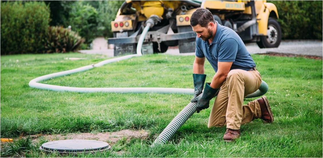 A worker kneels on a lawn, using a large hose to pump a septic tank connected to a yellow truck in the background.