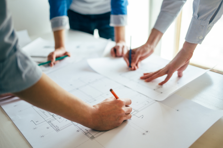 Hands and papers on a meeting table, with people sketching plans on a large sheet.