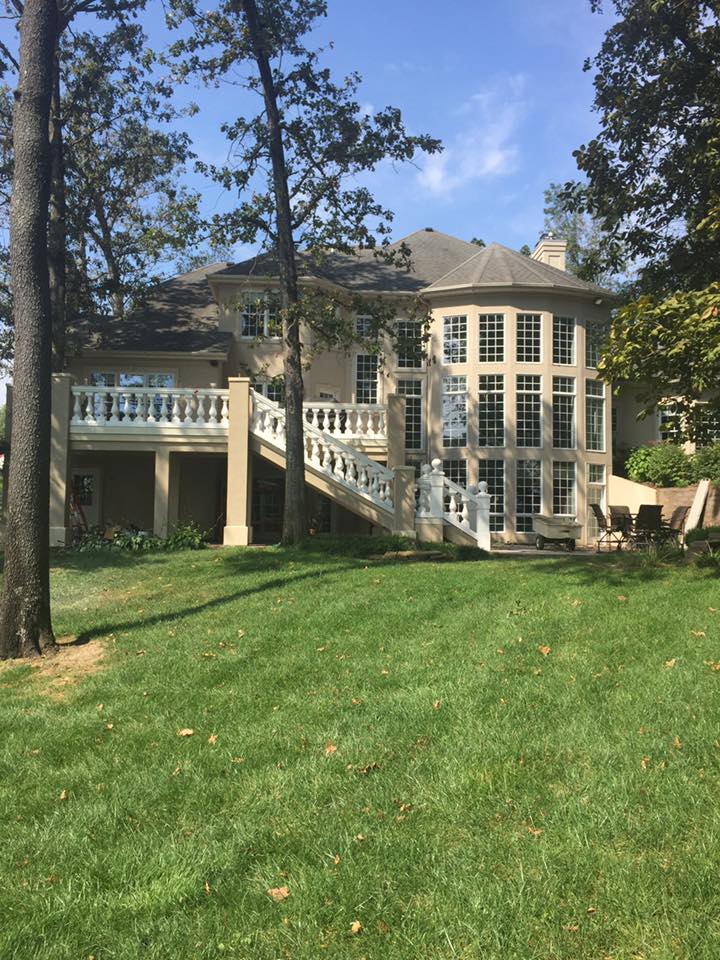Large two-story house with tall windows and white stairs, partly hidden by trees on a grassy lawn.